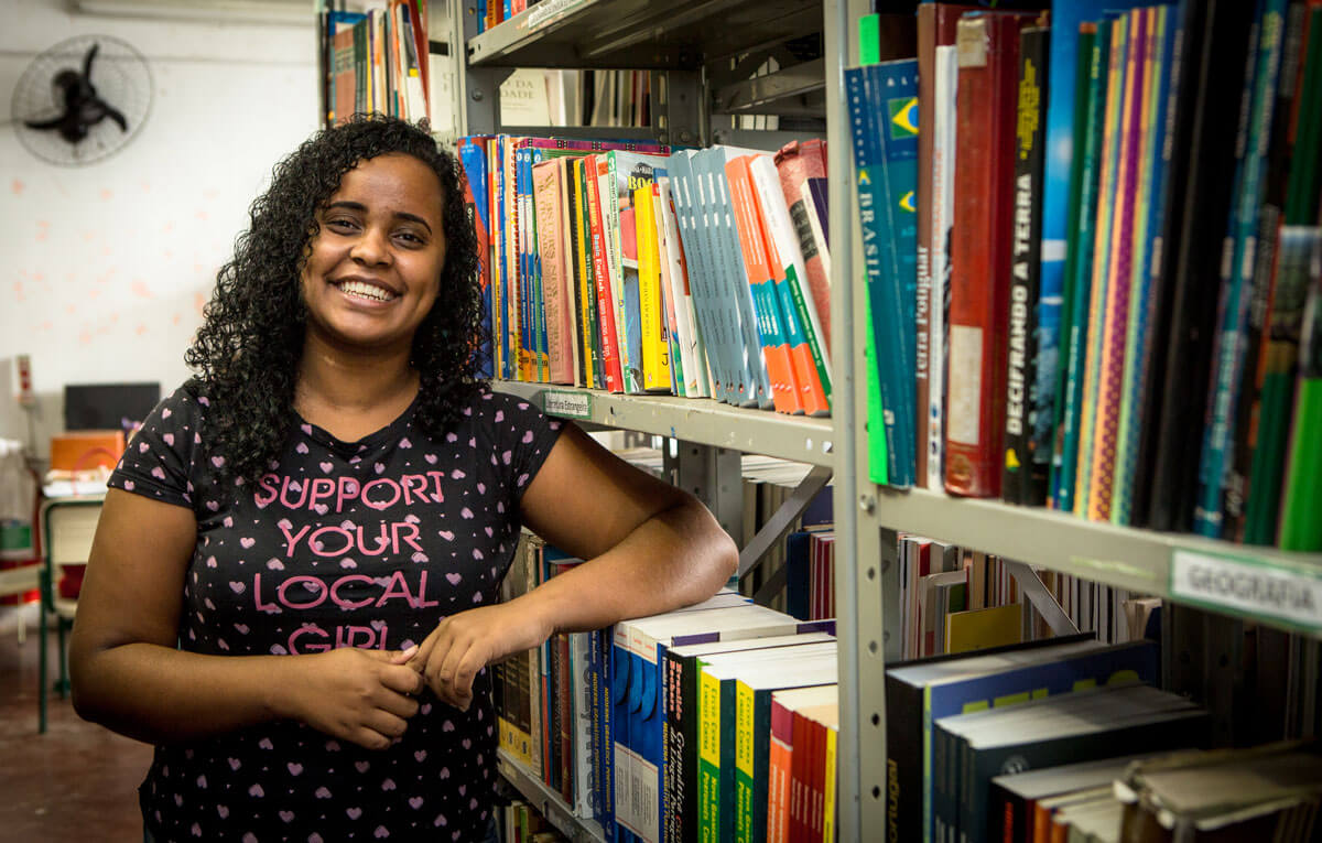 Ex-aluna Thaianne Santos sorrindo ao lado de uam estante de livros
